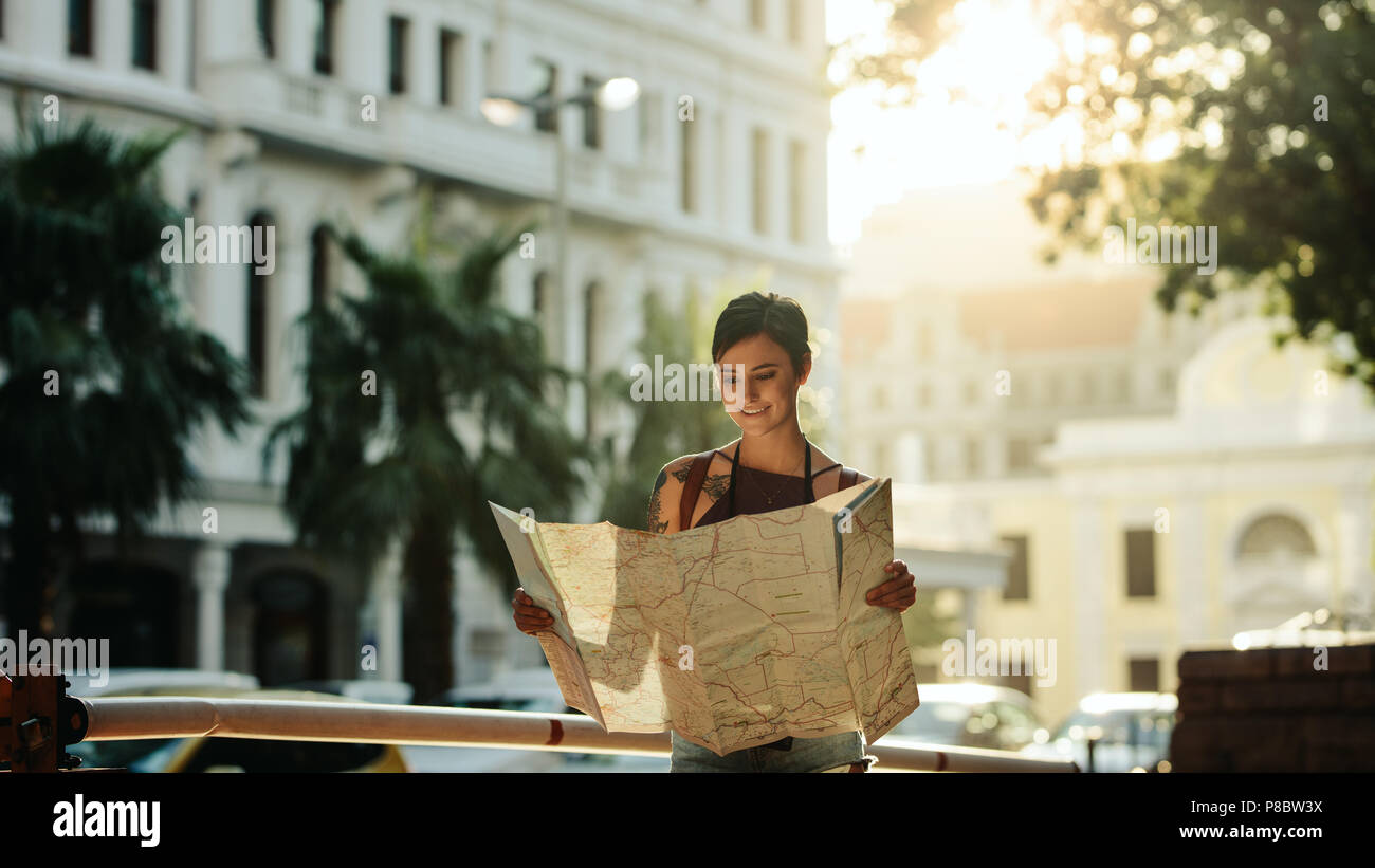 Touriste à l'aide d'une carte pour explorer la ville. Smiling woman standing on the street map avec des bâtiments de la ville en arrière-plan. Banque D'Images