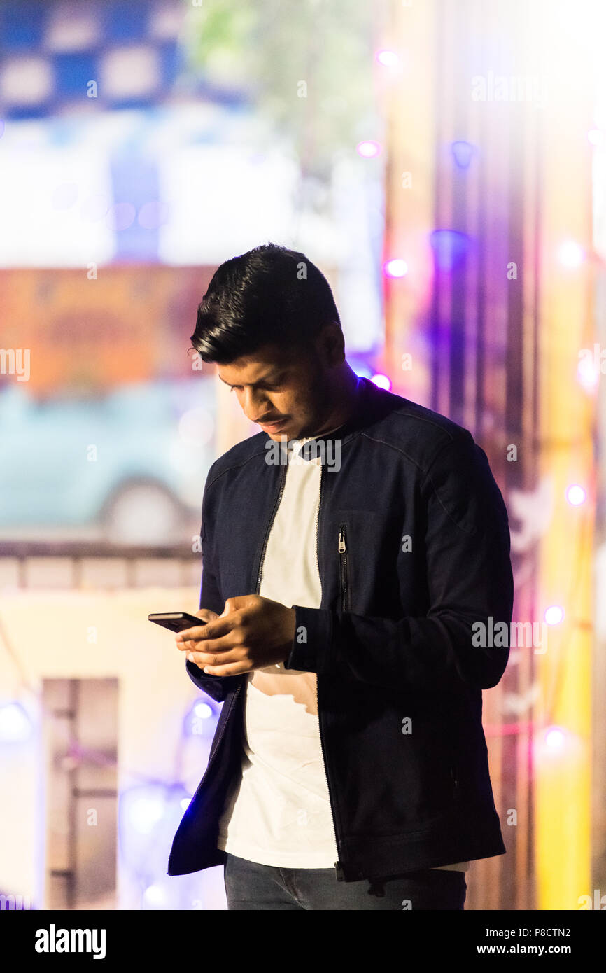 Jusqu'à la taille, portrait d'un jeune adulte jeune Indien en noir veste décontractée à l'aide de téléphone sur l'arrière-plan flou lumineux. Banque D'Images