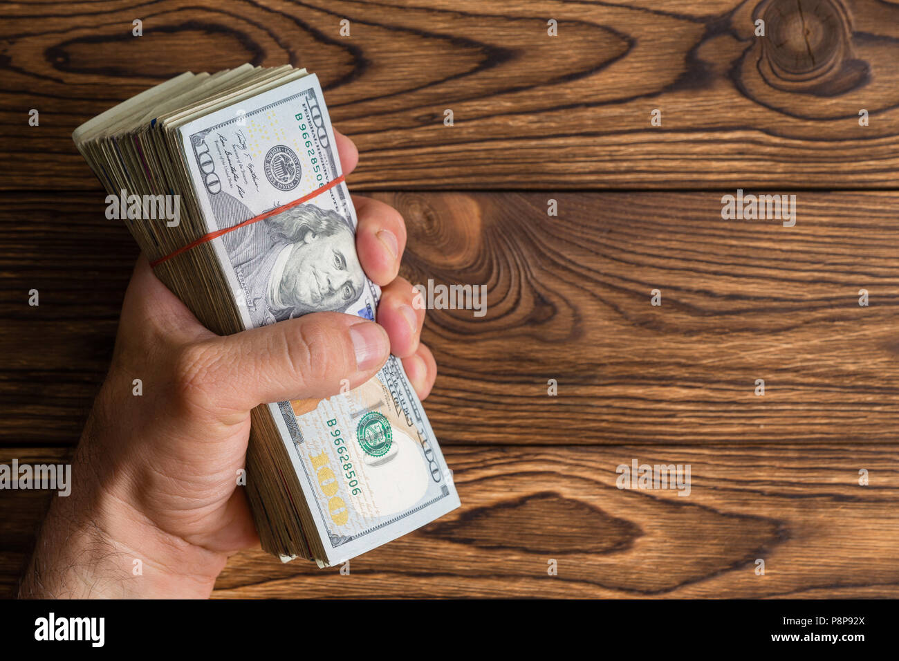 La préhension de l'homme une épaisse pile de 100 dollar bills ou Benjamins dans sa main sur un fond de bois rustique with copy space Banque D'Images