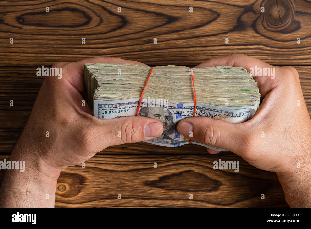 La préhension de l'homme une épaisse pile de 100 dollar bills ou Benjies fermement entre ses doigts sur une table en bois dans un cadre conceptuel de droit généraux Banque D'Images