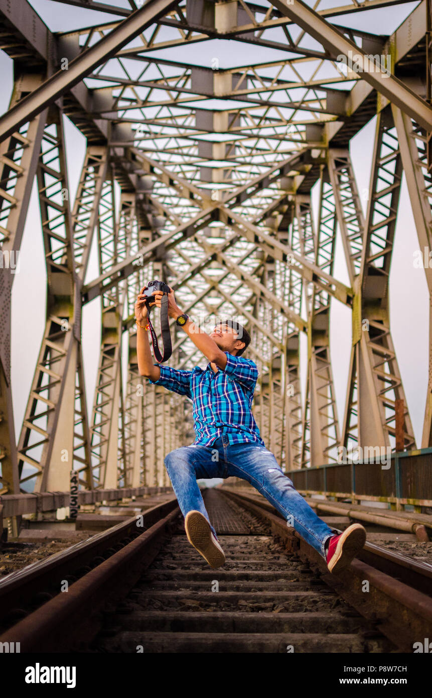 Jeune adulte Jeune Indien en tenant un appareil photo reflex avec selfies lévitation tandis que sur des rails de chemin de fer sous la structure du pont métallique symétriques. Banque D'Images
