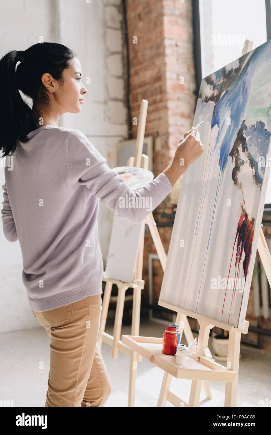 Side view portrait of young female artist holding peinture palette photos à chevalet dans art studio debout dans la lumière du soleil, copy space Banque D'Images