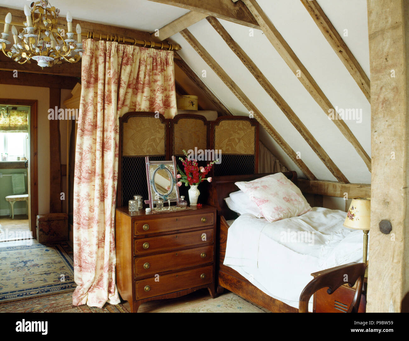 Une commode ancienne-de-tiroirs et l'écran à côté d'un lit simple dans une chambre mansardée avec une toile-de-Jouy à travers le rideau de porte Banque D'Images