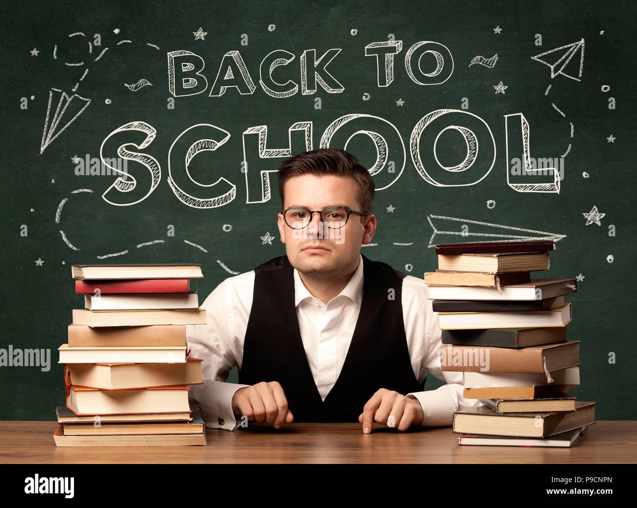 Un jeune enseignant dans les verres assis à classe 24 avec pile of books in front of blackboard disant concept dessin retour à l'école. Banque D'Images