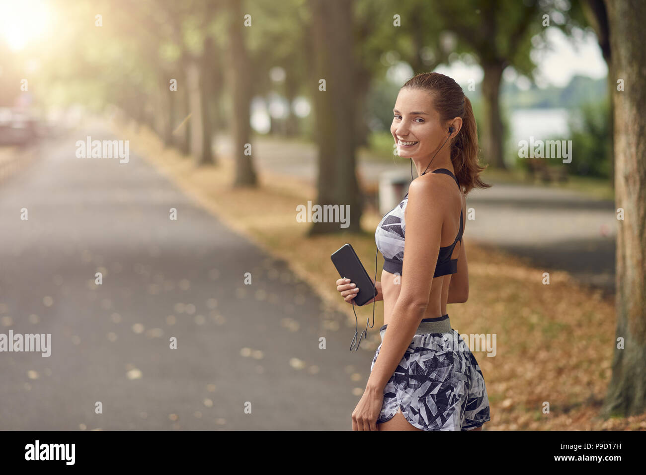 Jolie femme sportive à l'écoute de la musique sur son téléphone mobile sur le jogging sur une route bordée d'arbres tournant à sourire joyeusement à l'appareil photo Banque D'Images