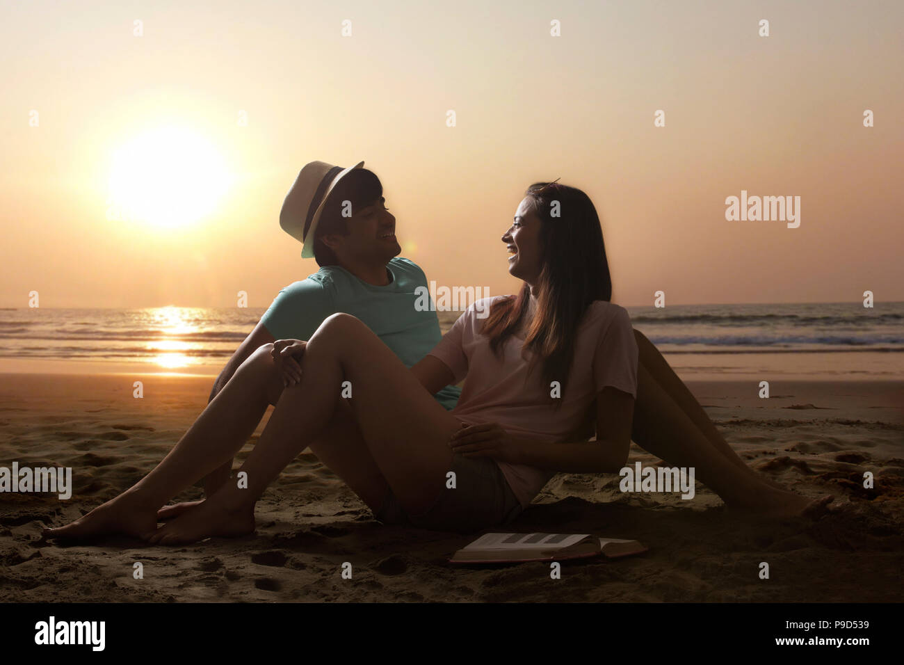 Smiling young couple sitting on beach at sunset Banque D'Images