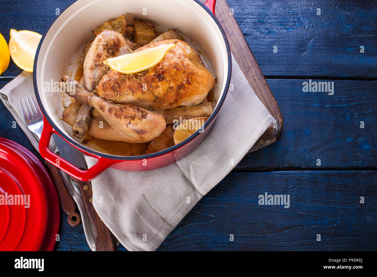 Le poulet entier cuit au four avec du citron et du romarin dans une fonte  rouge. Fond en bois bleu et gris serviette. Couteau et fourchette. Espace  libre pour le texte Photo