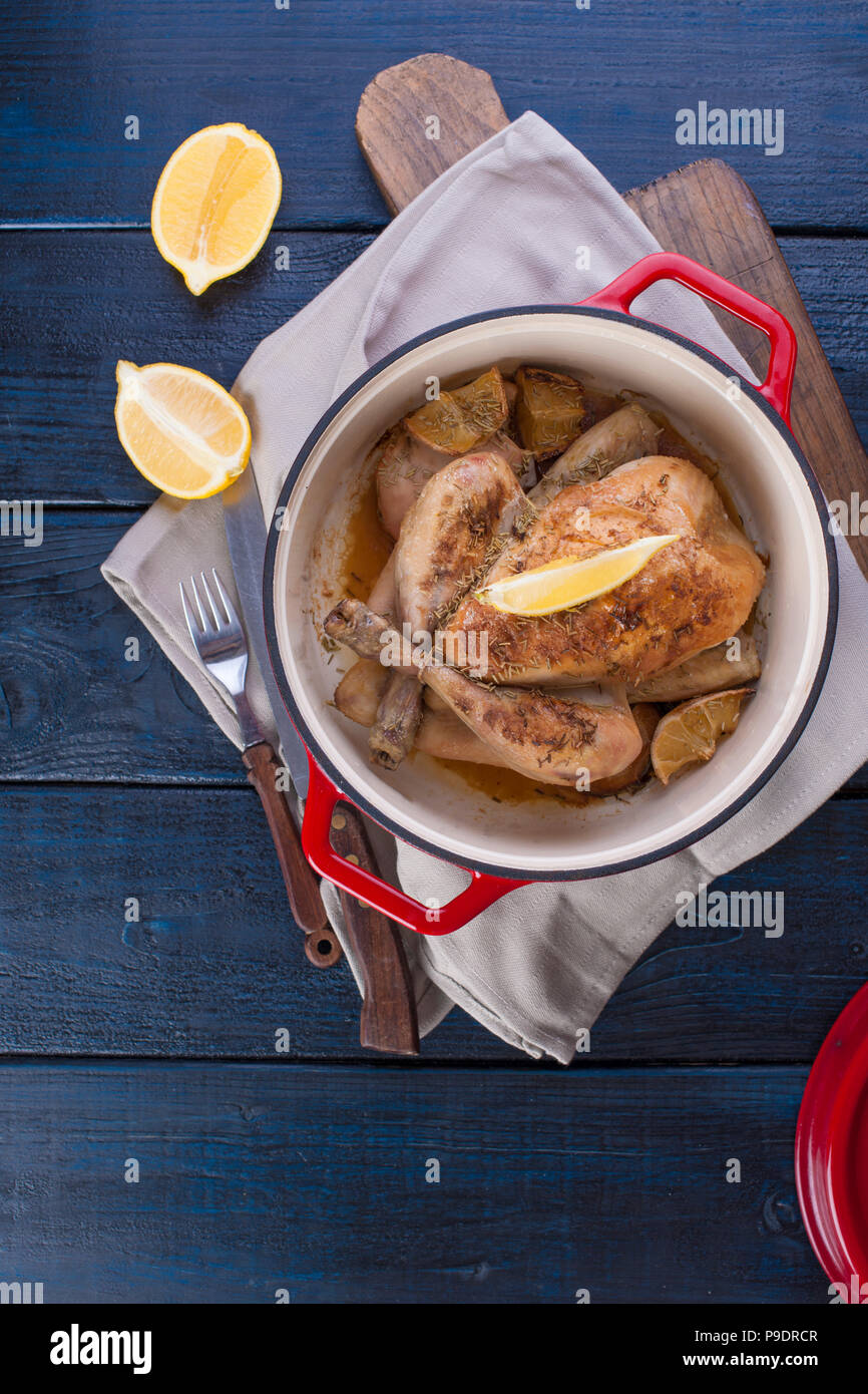 Le poulet entier cuit au four avec du citron et du romarin dans une fonte rouge. Fond en bois bleu et gris serviette. Couteau et fourchette. Espace libre pour le texte Banque D'Images