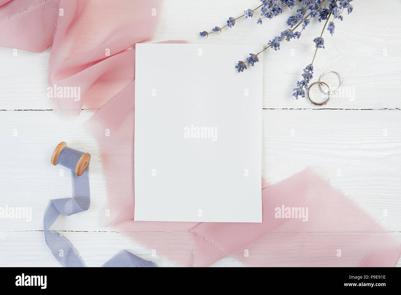 Carte vierge blanc ruban avec deux anneaux de mariage sur un fond de tissu rose avec des fleurs de lavande sur un fond blanc. Immersive avec enveloppe et carte vierge. Mise à plat. Vue d'en haut Banque D'Images