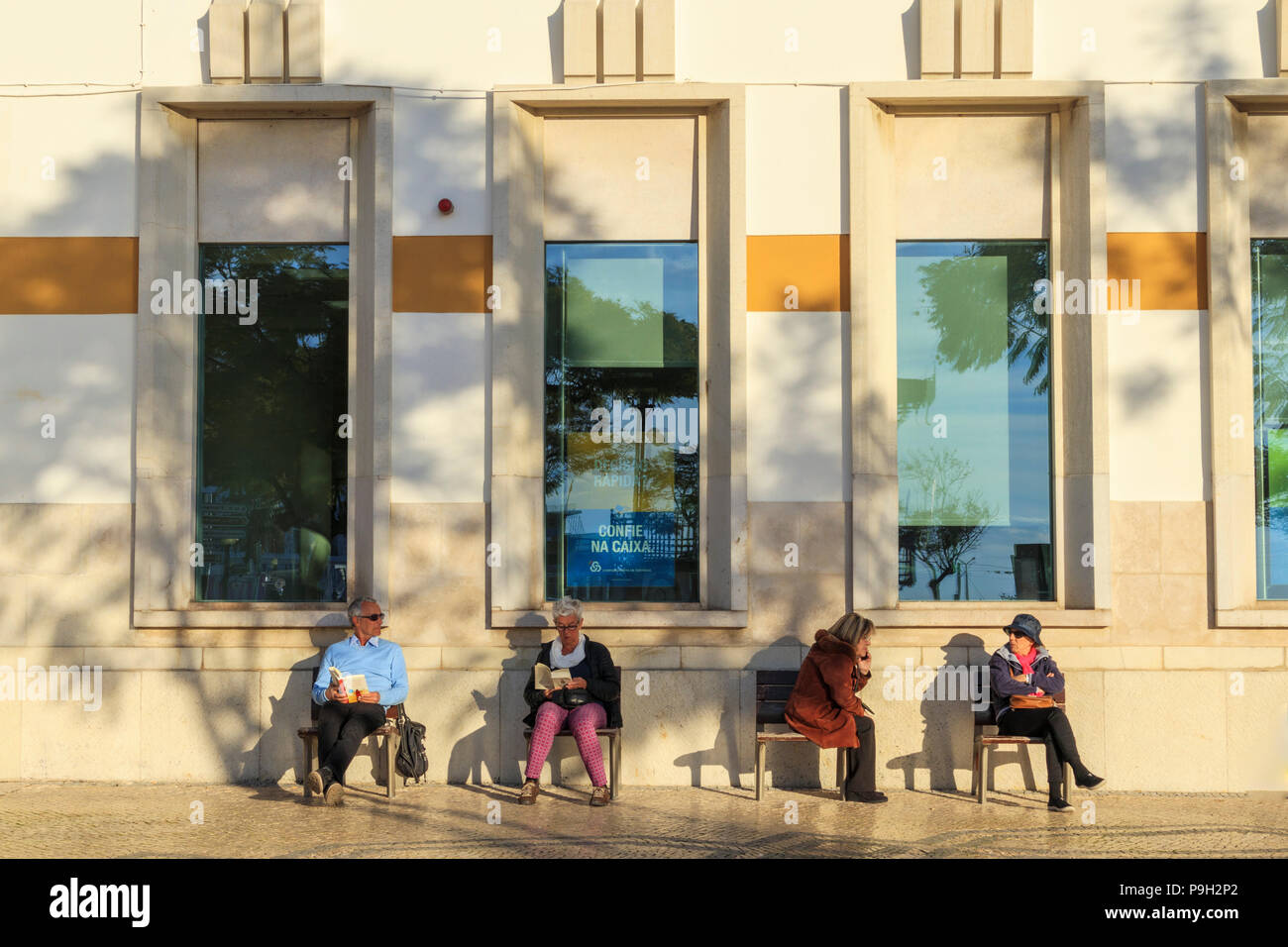Quatre personnes senior homme et femme sitrting dans le soleil du soir sur les sièges en bois placé à l'extérieur d'un grand bâtiment Faro Portugal Banque D'Images