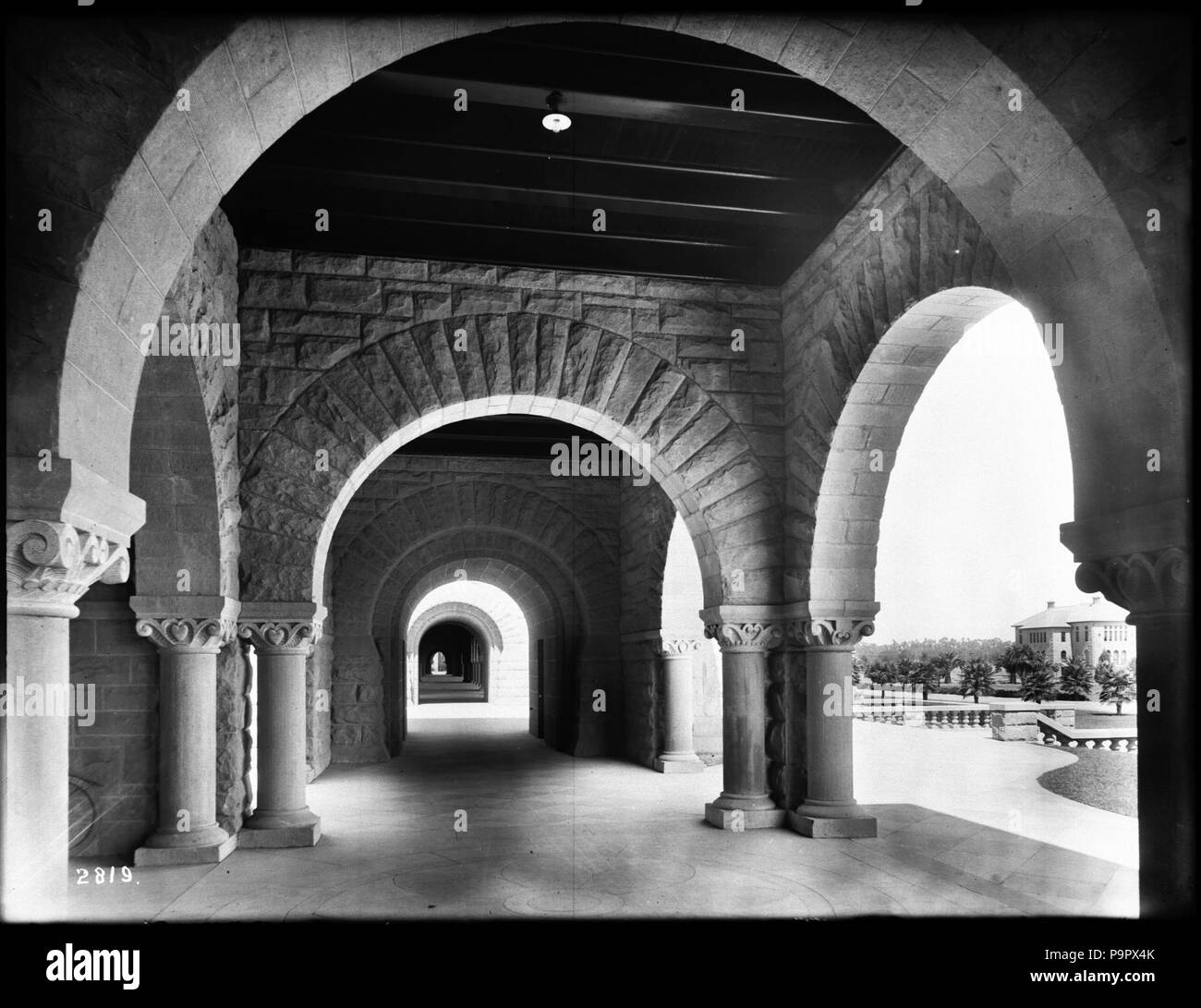 . Anglais : un corridor extérieur à l'Université de Stanford, à Palo Alto, Californie, ca.1900 Photo d'un corridor extérieur à l'Université de Stanford, à Palo Alto, Californie, ca.1900. Au centre, un grand couloir peut être vu s'étendant dans le contexte extrême. Arches de pierre de taille d'encadrer le couloir. À la base de chaque roue, une colonne décorative en pierre peut être vu. Au-dessus du couloir, de grandes poutres de soutien et d'un seul appareil d'éclairage sont visibles. À droite à travers l'une des arches, un bâtiment et une partie du paysage sont visibles. Plusieurs grands palmiers peut être vu à côté de l'immeuble. Banque D'Images