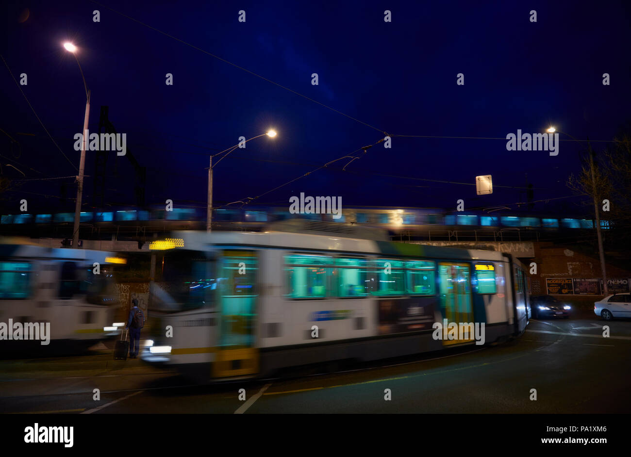 Un homme est toujours en attente d'un tram comme deux tramways passent dans les deux sens devant et derrière lui. Un train passe sur le pont juste au-dessus. Banque D'Images