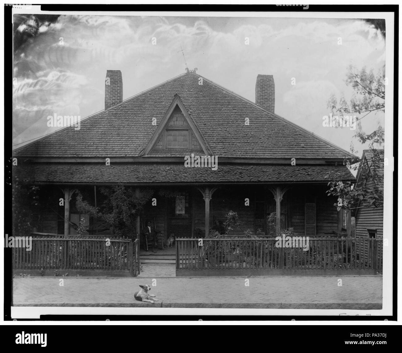 634 Vue extérieure de la maison avec des piquets de clôture, l'homme et le chien assis sur le porche et un chien couché sur le trottoir RCAC99472382 Banque D'Images