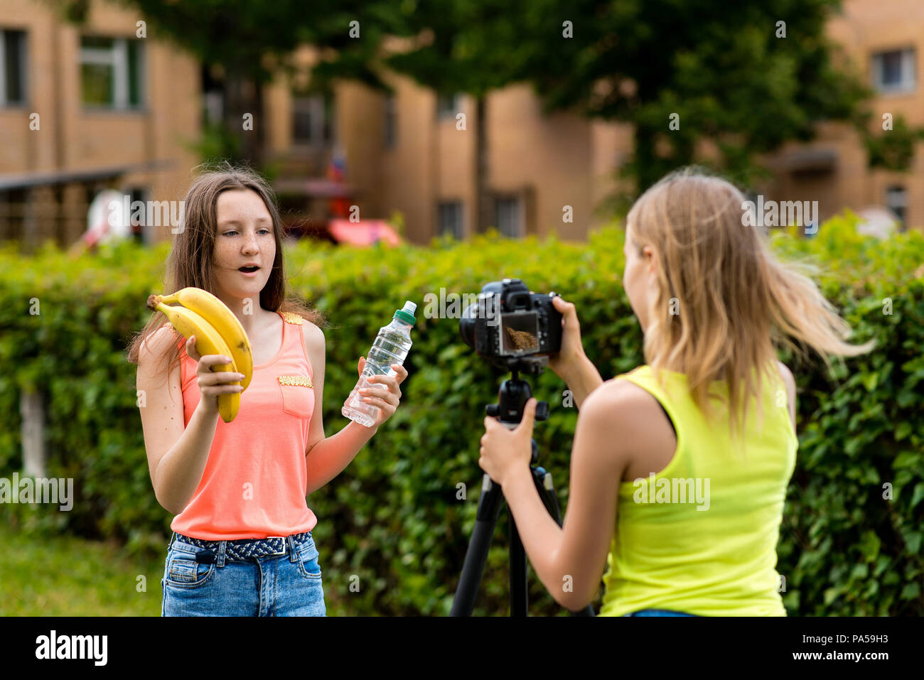 Les jeunes filles des blogueurs, à l'été dans la ville à la nature. Enregistrer une vidéo sur l'appareil photo. Dans ses mains tient une bouteille d'eau et deux bananes. Le conce Banque D'Images
