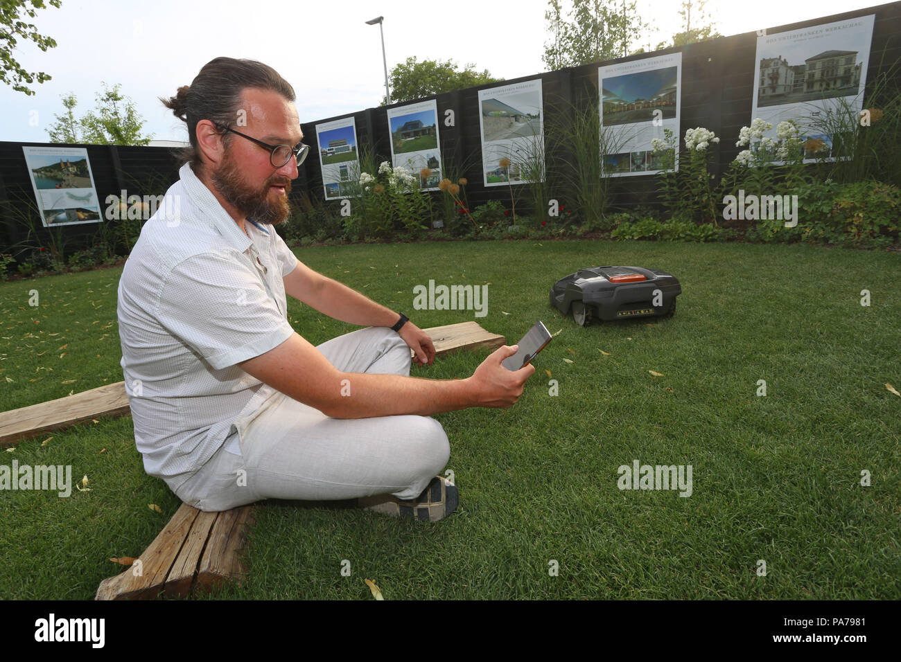Würzburg, Allemagne. Le 04 juillet, 2018. Alexander Seufert contrôlant un robot lawnmowing avec son smartphone dans le 'jardin' de gagner du temps dans le jardin de l'État de Bavière Show. Le jardin conçu par ce maître montre que le jardinage jardin numérique n'est pas une chose de l'avenir. Credit : Karl-Josef Opim/dpa/Alamy Live News Banque D'Images