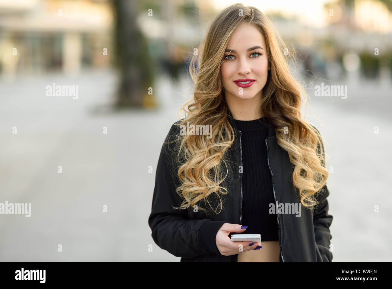 Blonde woman texting avec son smart phone en contexte urbain. Belle jeune fille portant veste noire marchant dans la rue. Jolie femme russe Banque D'Images