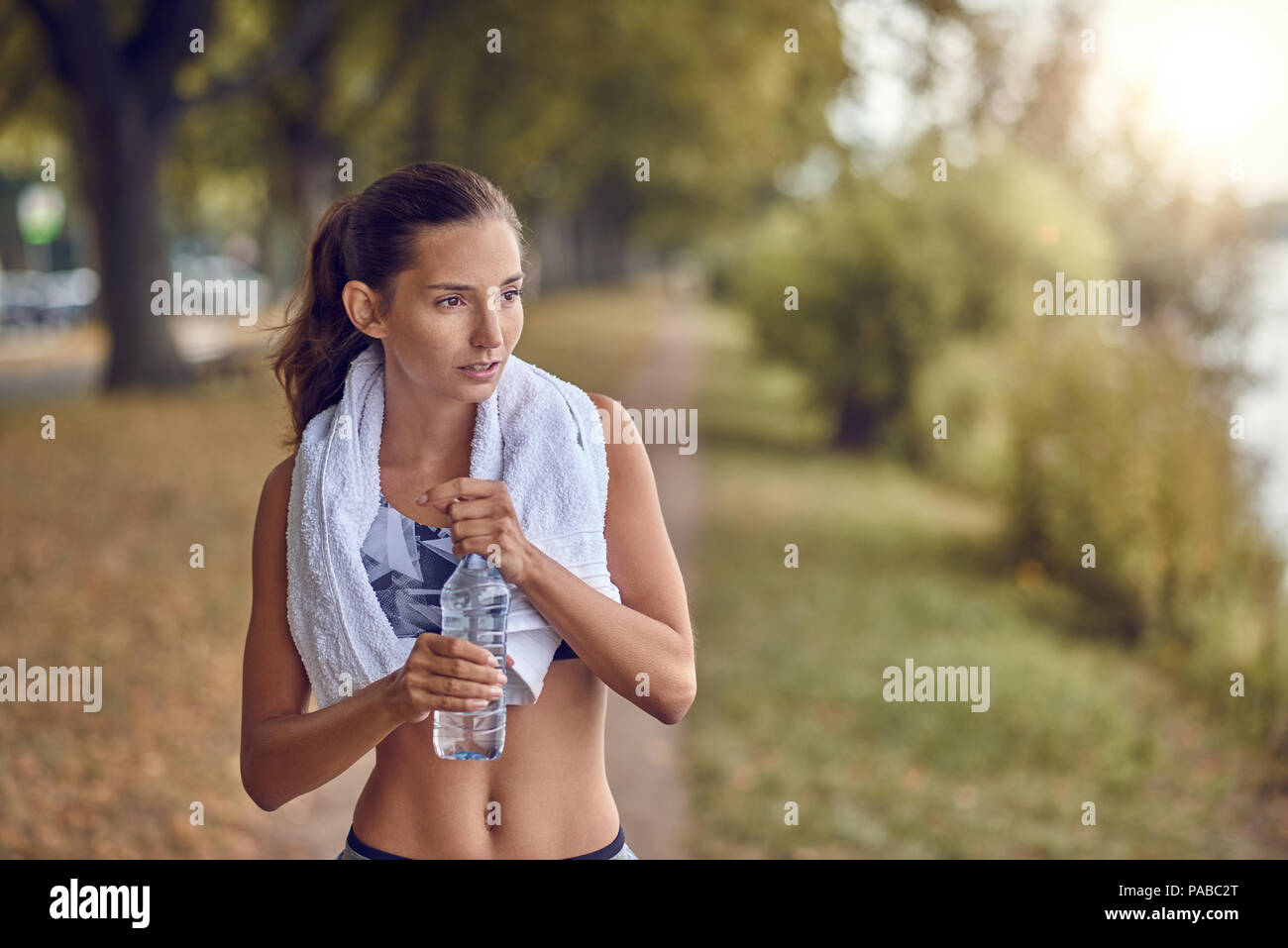 Mettre en place les plus sportifs femme tenant une bouteille d'eau qu'elle fait une pause au cours de son jogging entraînement sur un sentier bordé d'arbres avec une serviette autour du cou Banque D'Images