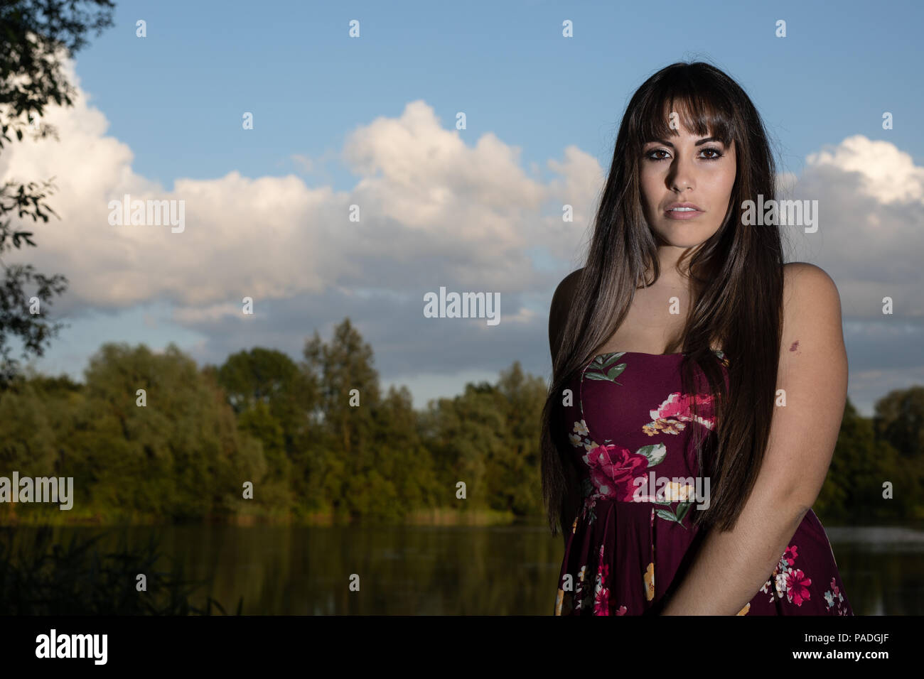 L'Amérique espagnole femme debout au bord d'un lac dans le Cambridgeshire. Portant une robe couleur prune lors d'un coucher du soleil. Banque D'Images