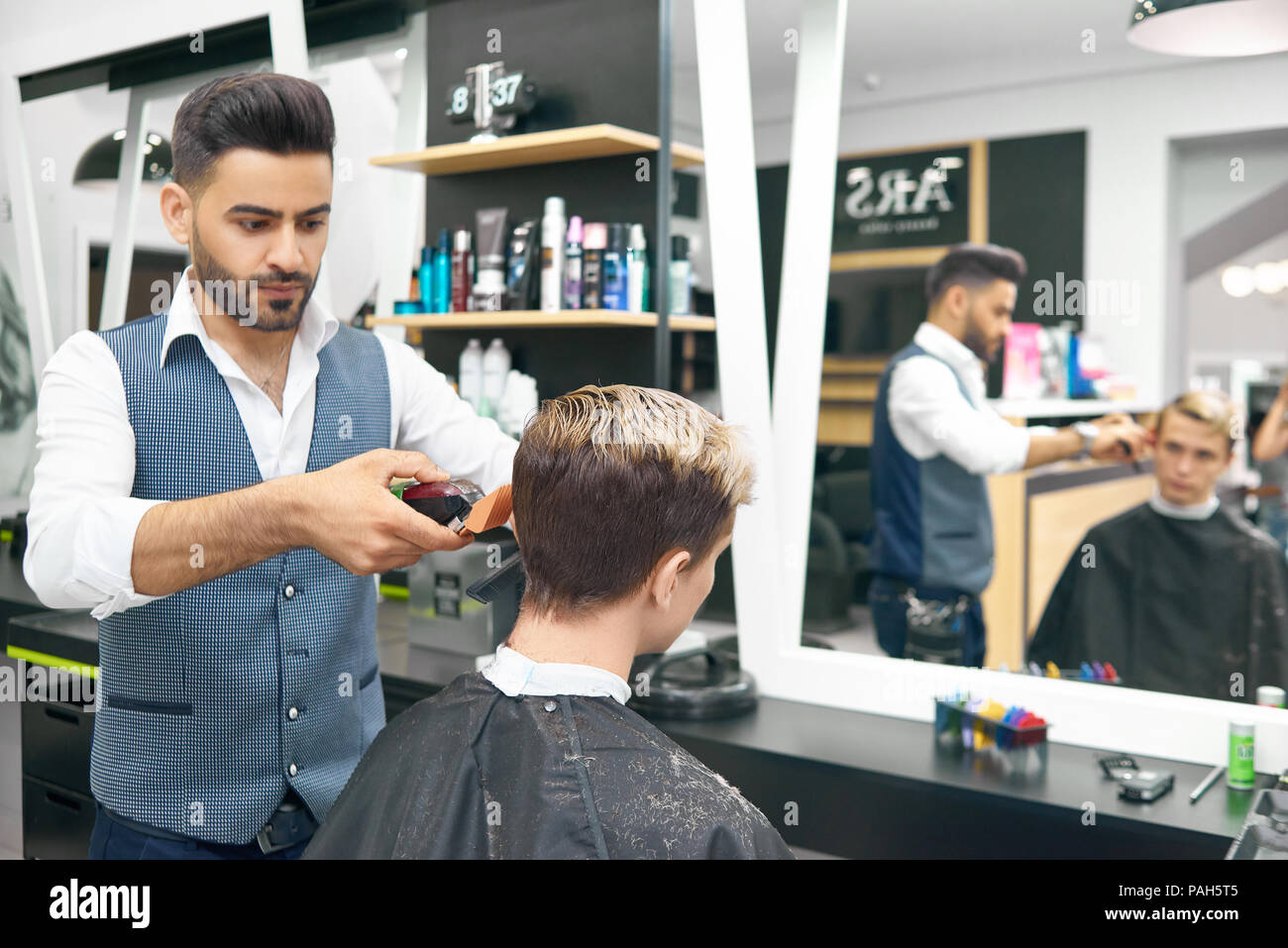 Faire nouvelle coiffure moderne pour jeune homme dans un salon de coiffure. L'utilisation de clipper, hairstylist professionnel à concentré. Modèle masculin assis devant un grand miroir, recouvert d'une cape noire. Banque D'Images