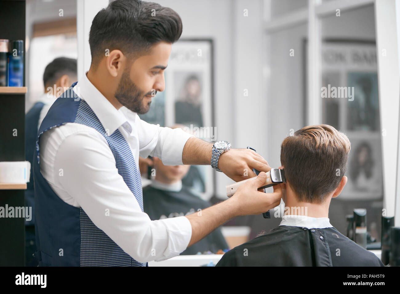 Salon de coiffure faisant nouvelle coupe pour young client assis devant un miroir. Vêtu de blanc chemise décontractée, gilet gris, regardez. À la recherche d'amour, concentré son travail. Modèle recouvert d'une cape noire. Banque D'Images