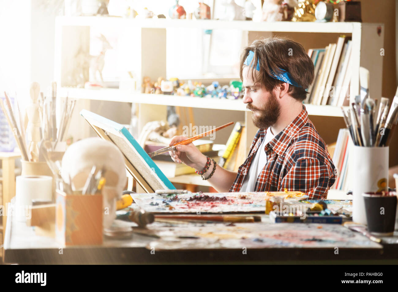 Jeune homme de race blanche à l'artiste avec dessin bandana bleu brosse et huile sur toile dans son studio ensoleillé Banque D'Images