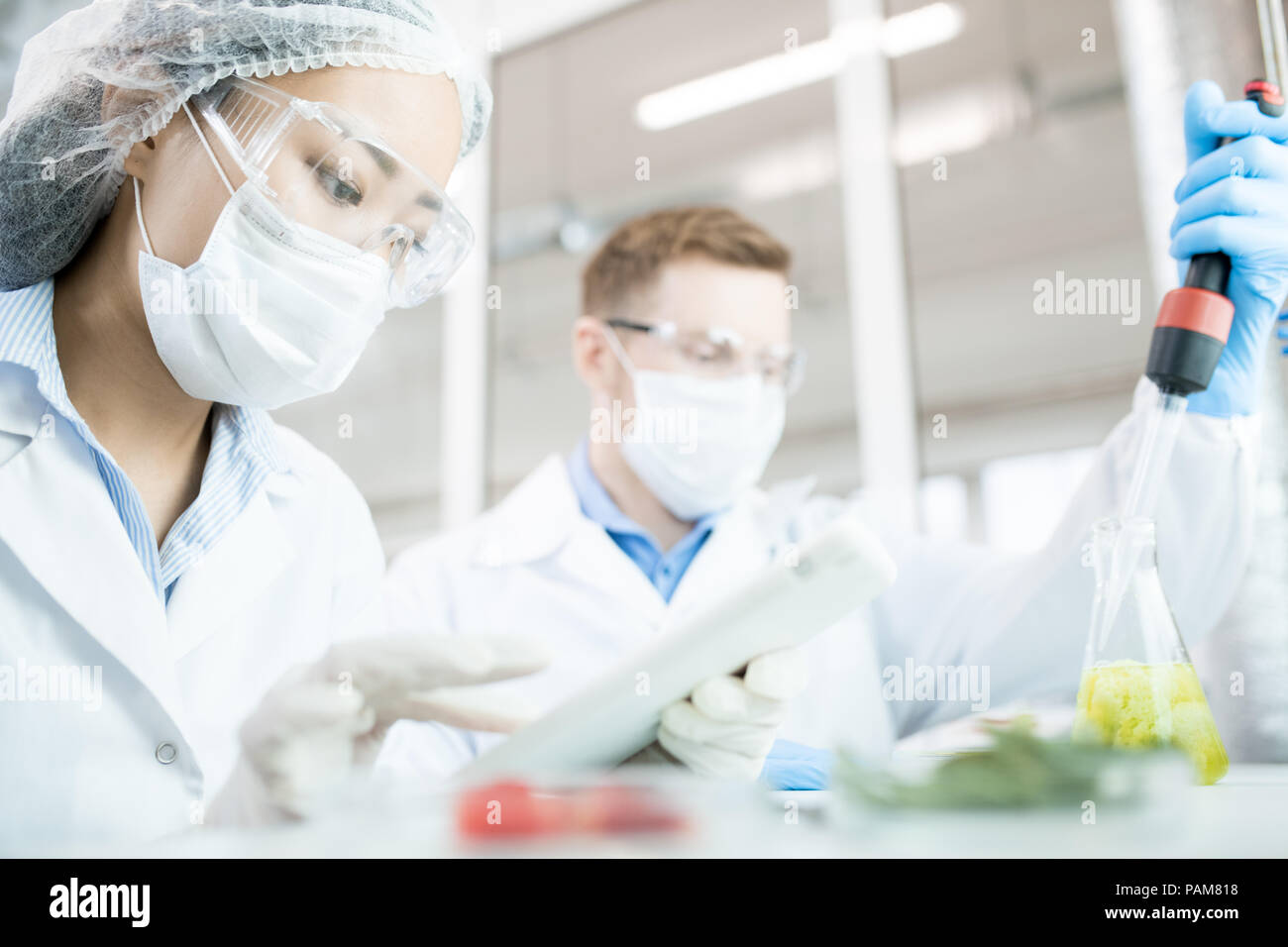 Portrait de deux scientifiques modernes portant des masques qui travaillent sur l'étude de recherche sur les aliments liquides dans le bécher tout en sitting at table in laboratory Banque D'Images