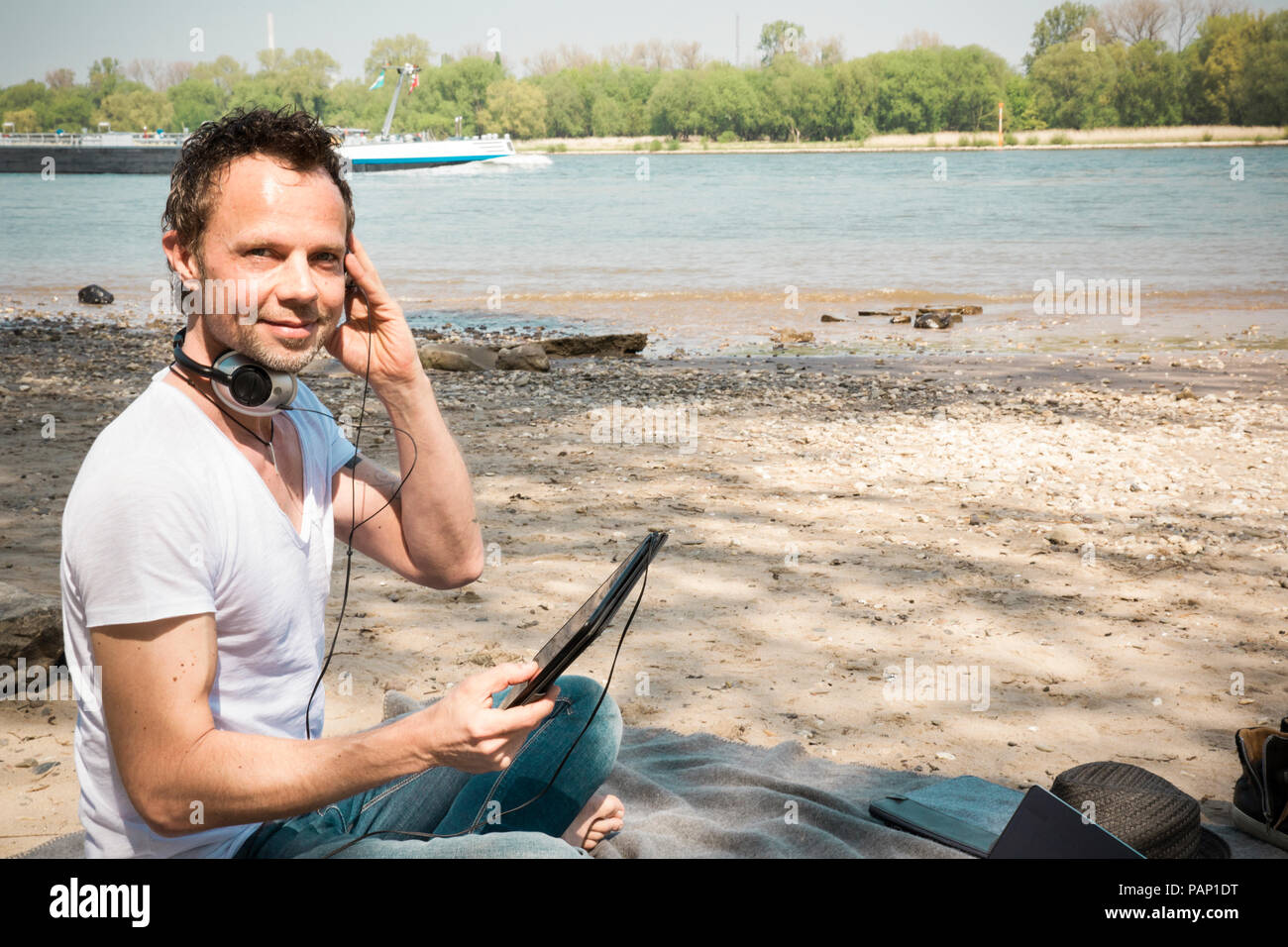 Portrait of smiling man sitting on couverture à une rivière à l'aide de tablet Banque D'Images