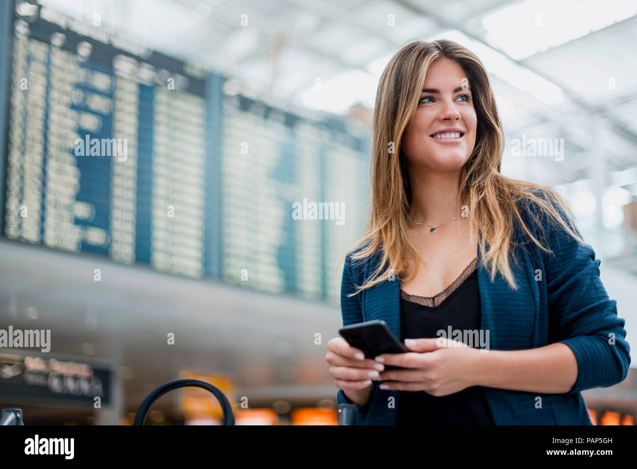 Smiling young woman with cell phone at departure board à travers Banque D'Images