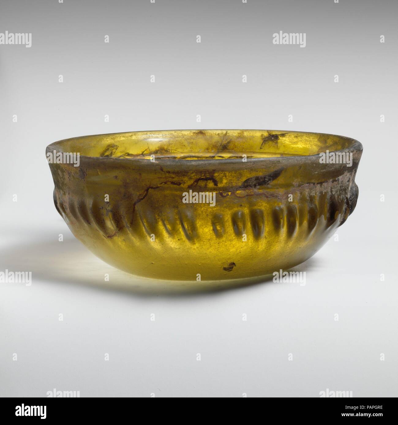 Bol côtelé en verre. Culture : le grec, l'Est de la Méditerranée. Dimensions : H. : 2 3/16 in. (5,6 cm) Diam. : 5 3/8 in. (13,7 cm). Date : premier siècle avant J.-C.. Jaune Vert translucide. Outsplayed rim, avec bord arrondi et lisse vers l'intérieur de la pente au-dessous de la bande ; face convexe de la courbe vers le bas ; fond concave. Sur l'intérieur, une seule large rainure horizontale à la jonction de la jante et le côté ; à l'extérieur, quarante-trois courtes, unevely-côtes espacées de différentes longueur et largeur, disposés autour de la section médiane renflée du corps, à l'occasion, flanquée par vertical des marques d'outillage. Intacts ; certaines tensions internes ; certains Banque D'Images