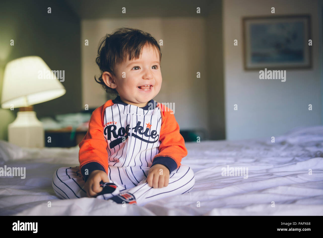 Portrait of laughing baby girl wearing jumpsuit sitting on bed Banque D'Images