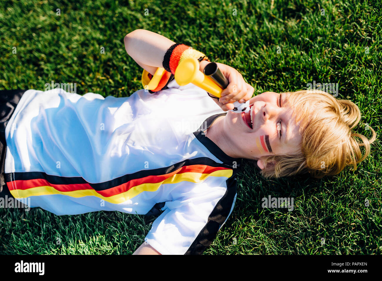 Garçon en allemand soccer shirt lying on grass, blowing horns Banque D'Images