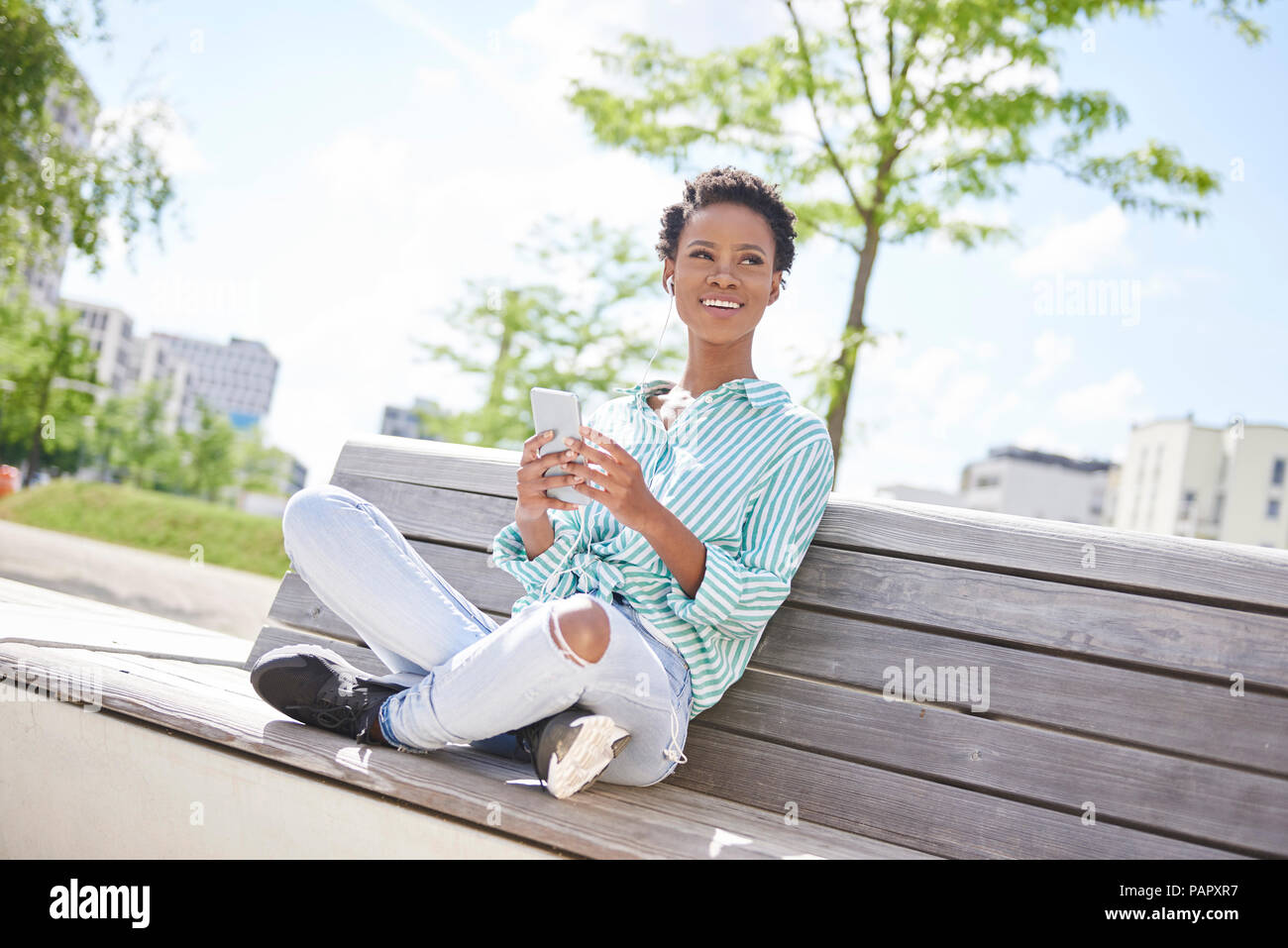 Portrait of smiling young woman with cell phone et écouteurs assis sur un banc Banque D'Images