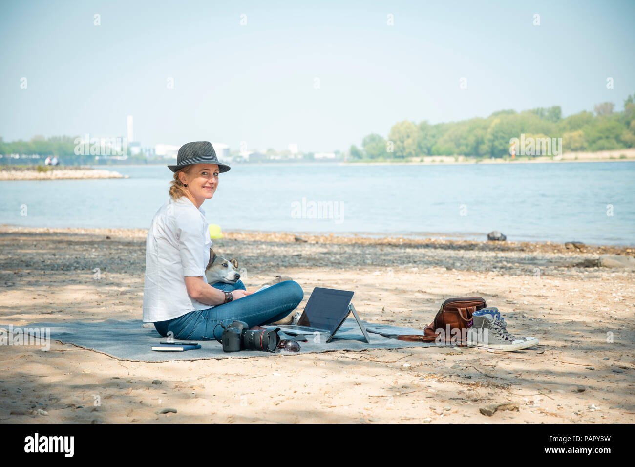 Smiling woman sitting on couverture à une rivière avec chien et coffre Banque D'Images