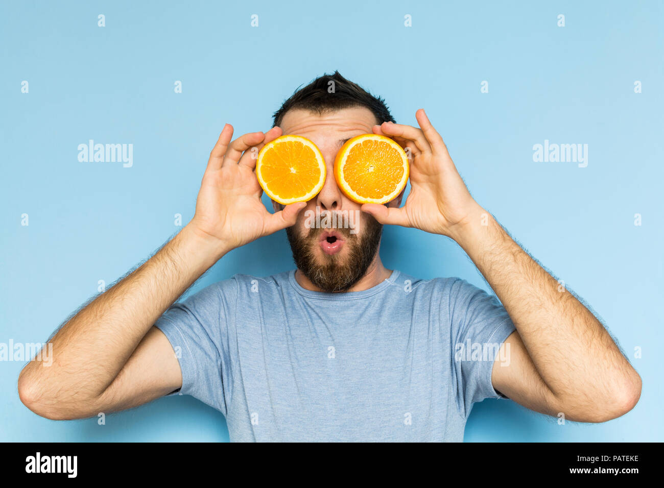 Jeune homme barbu tenant tranches d'orange en face de ses yeux. Fond bleu clair, expression surprise sur le visage de l'homme. Banque D'Images