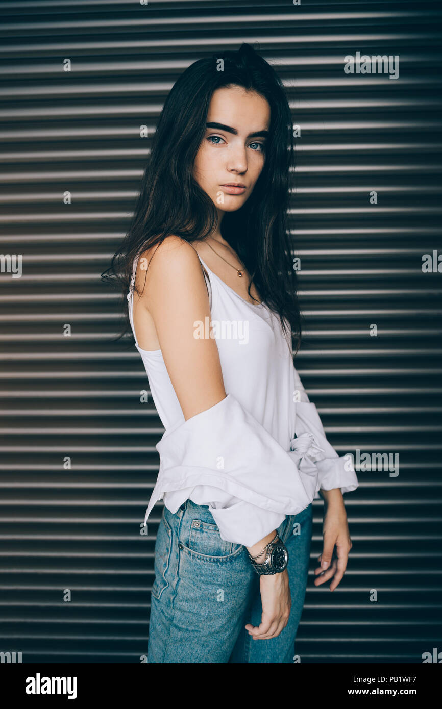 Belle jeune femme portant haut blanc abaissé sa veste pour montrer ce qui pose l'épaule à fond brun. Fille avec de longs cheveux noirs à l'extérieur permanent loo Banque D'Images