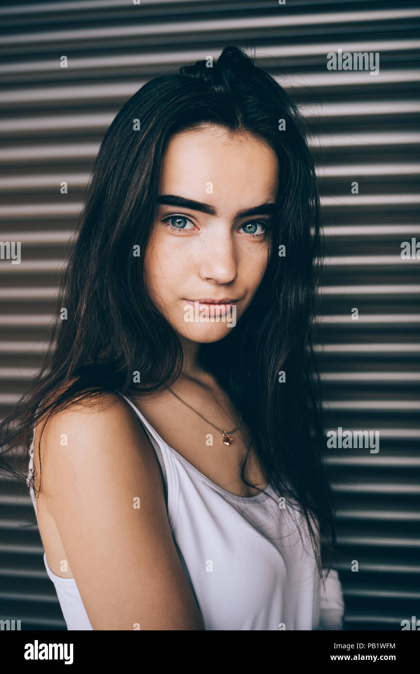 Portrait de jolie fille avec de longs cheveux noirs et les yeux bleus. Young woman wearing white top debout sur fond brun à la caméra et au sourire. Banque D'Images