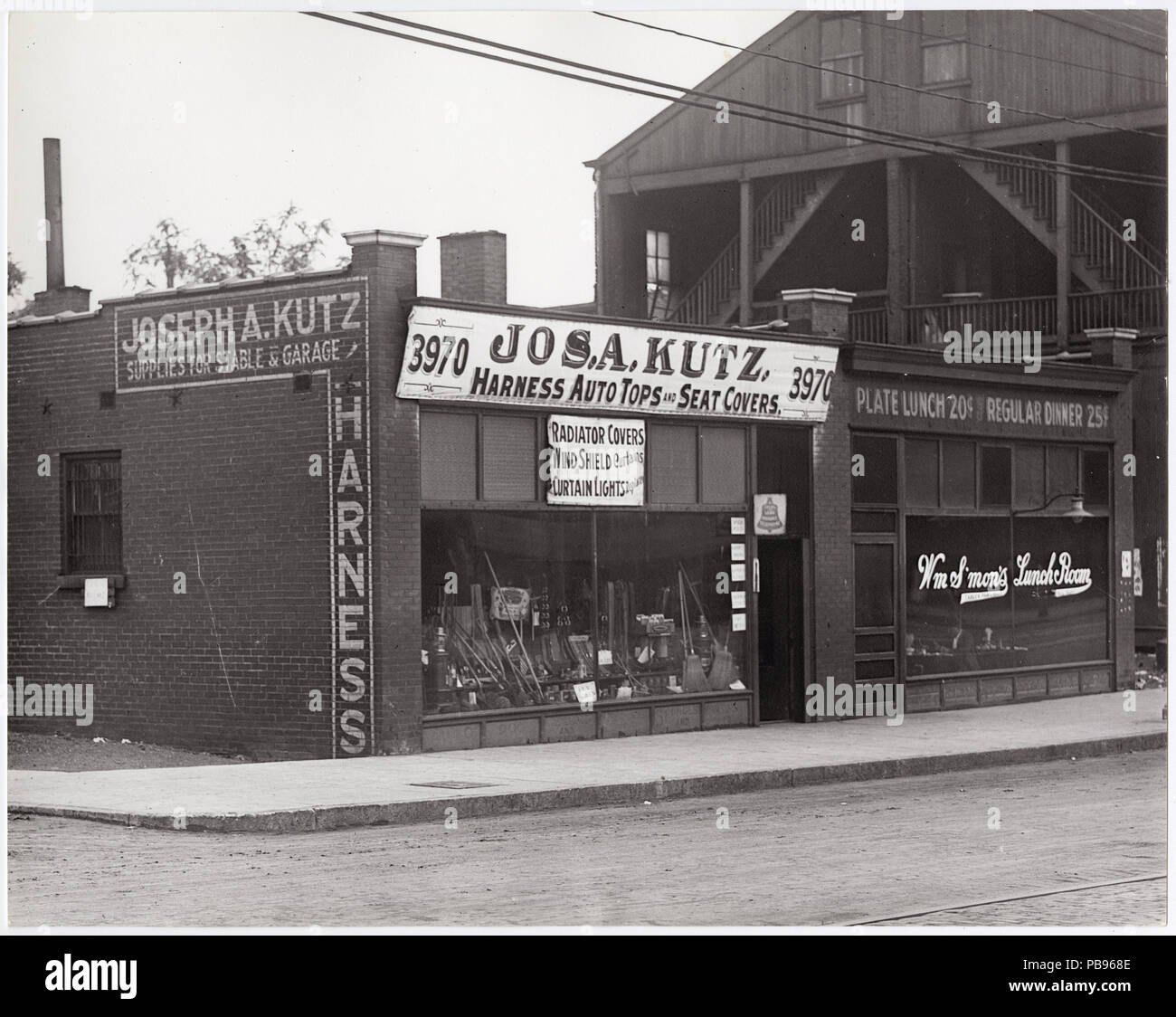 . Anglais : horizontale, noir et blanc photographie montrant Joseph A. Kutz's store au 3970 Chouteau. Le bâtiment en brique d'un étage dispose d'une grande fenêtre avant l'affichage des balais, des râteaux ou des fourches, de lampes, et d'autres équipements ont besoin d'une voiture ou stable. L'avant de signer le texte dit : "Jos. A. Kutz. Faisceau / Auto-tops et des housses de siège. Couvre Radiateur / / / rideaux pare-brise feux Rideau remplacé.' un signe peint sur le côté de l'édifice se lit comme suit : "Joseph A. Kutz / Fournitures pour stable et Garage / faisceau." La publicité des signes d'affaires voisins lire : 'HM S'mon's Lunch / Déjeuner Chambre plaque 20 cents / R Banque D'Images