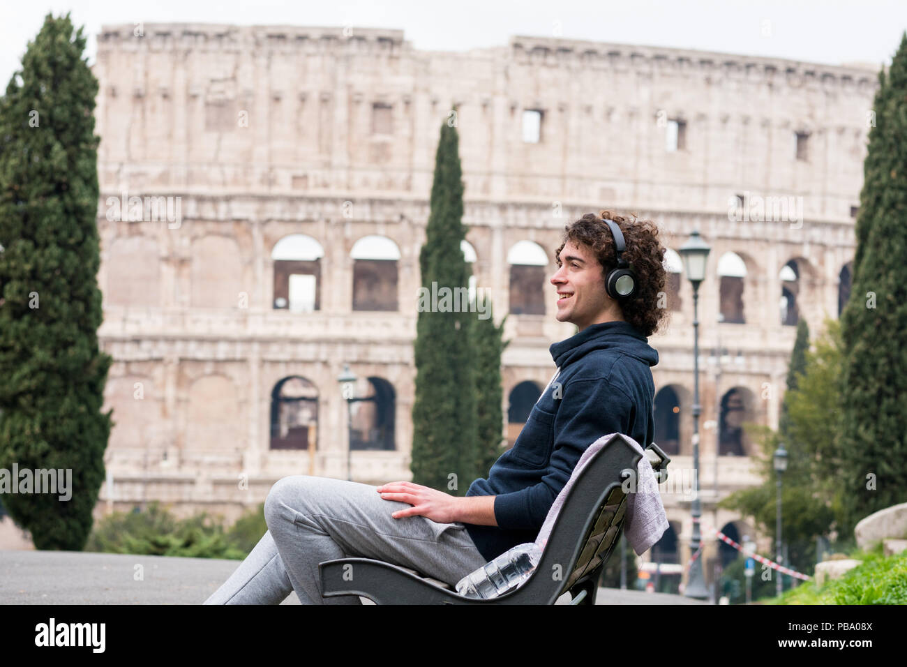 Beau jeune homme en chiffons sport écouter de la musique assis sur un banc avec le Colisée en arrière-plan. Les jeunes sportifs en survêtement 23-08-2003 détente Banque D'Images