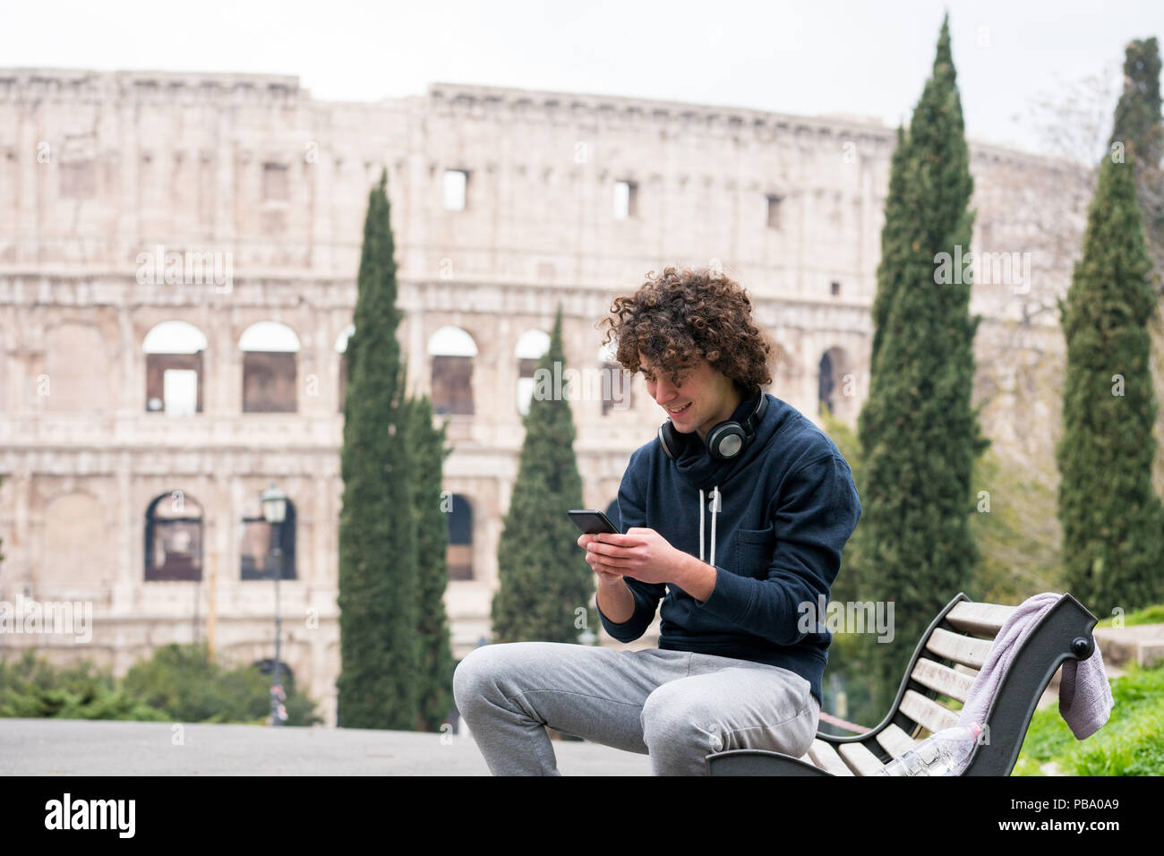 Beau jeune homme dans les sports à l'aide de chiffons de son téléphone mobile tout en restant assis sur un banc à Rome. Les jeunes sportifs en survêtement se détendre après la formation avec Banque D'Images