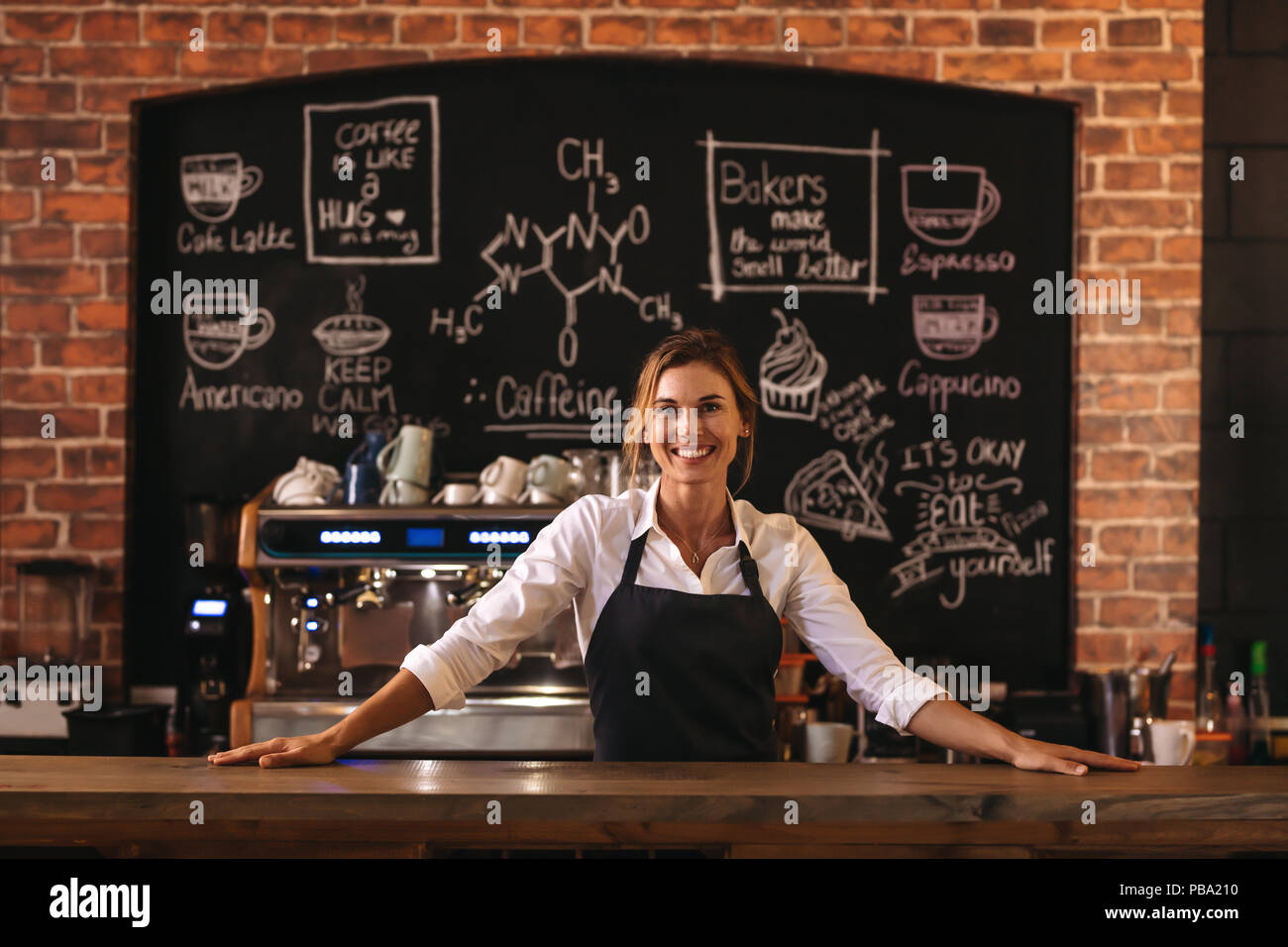 Portrait of female barista debout derrière comptoir. Femme cafe owner in apron looking at camera et souriant. Banque D'Images