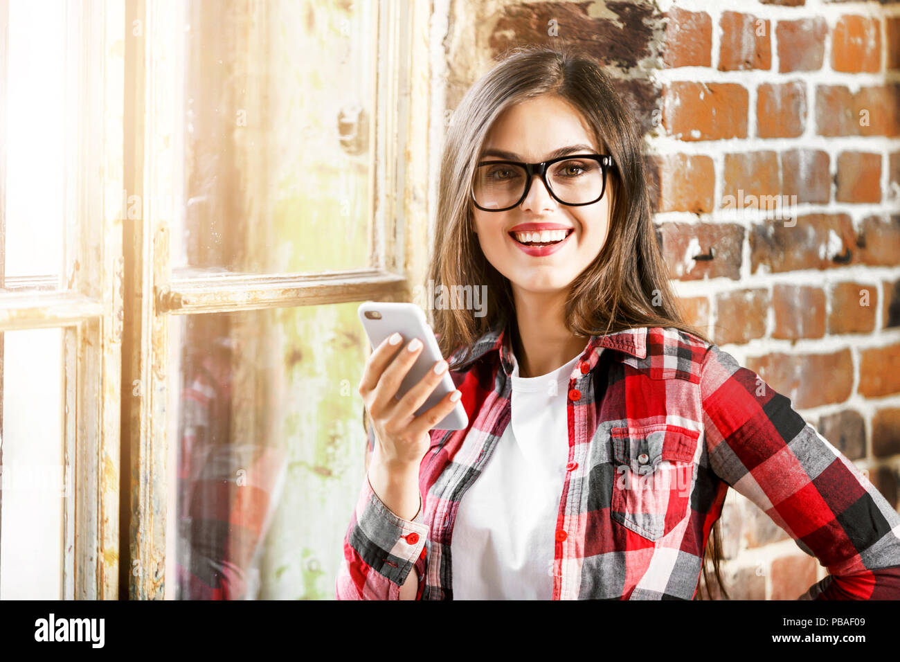 L'adolescent aux cheveux longs mignon femelle fille dans les verres à l'aide de téléphone mobile et souriant près de la fenêtre de la mezzanine bureau moderne Banque D'Images