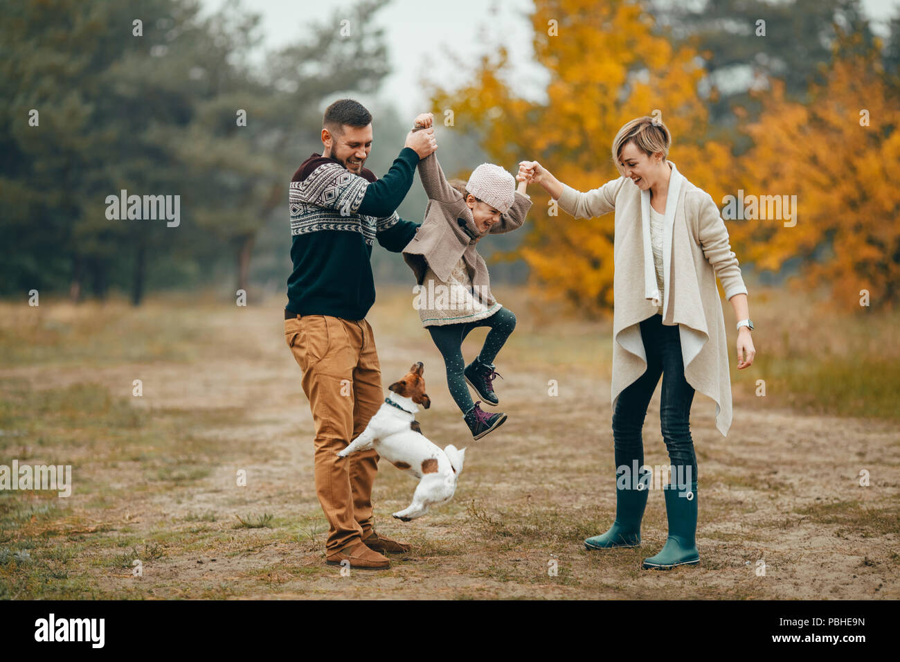Heureux parents s'amuser et élèvent leur fille au chemin forestier en regard de chien sautant au cours de marche en forêt d'automne. Banque D'Images
