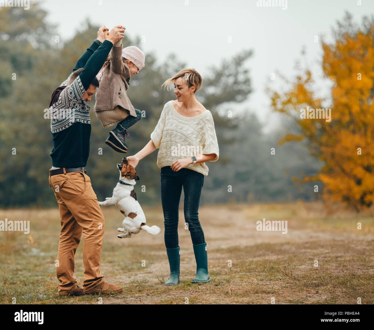 Heureux parents s'amuser et élèvent leur fille au chemin forestier en regard de chien sautant au cours de marche en forêt d'automne. Banque D'Images