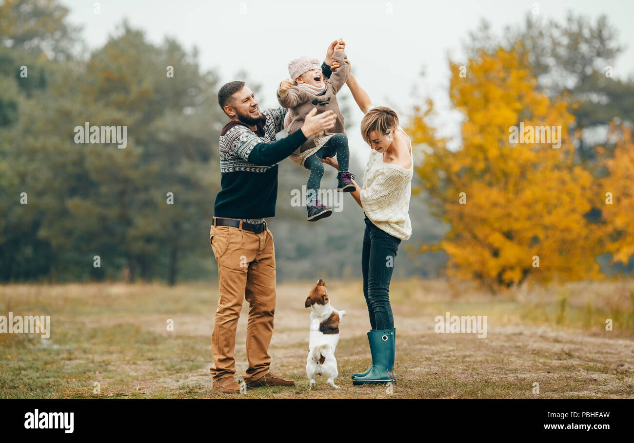 Heureux parents s'amuser et élèvent leur fille au chemin forestier en regard de chien sautant au cours de marche en forêt d'automne. Banque D'Images