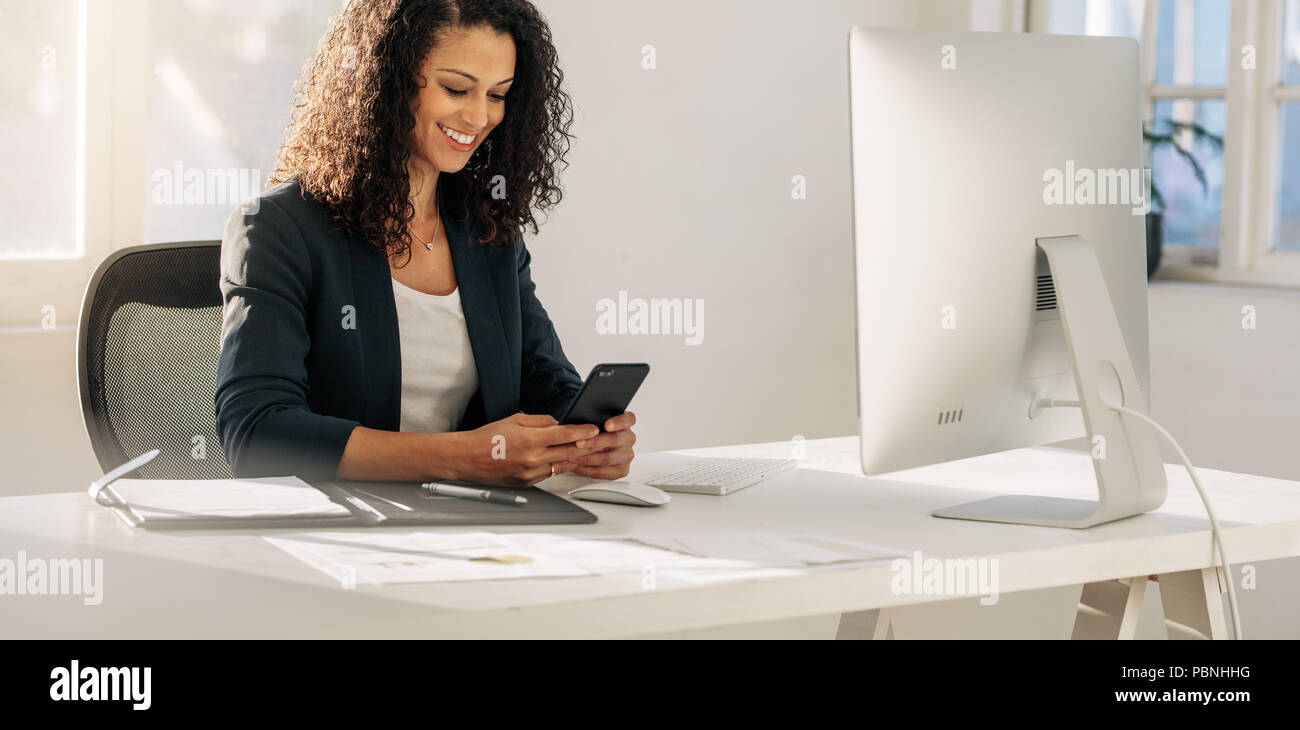 Smiling woman entrepreneur à son bureau dans son bureau de contrôle de téléphone mobile. Femme assise en face de l'ordinateur à la recherche de téléphone cellulaire. Banque D'Images