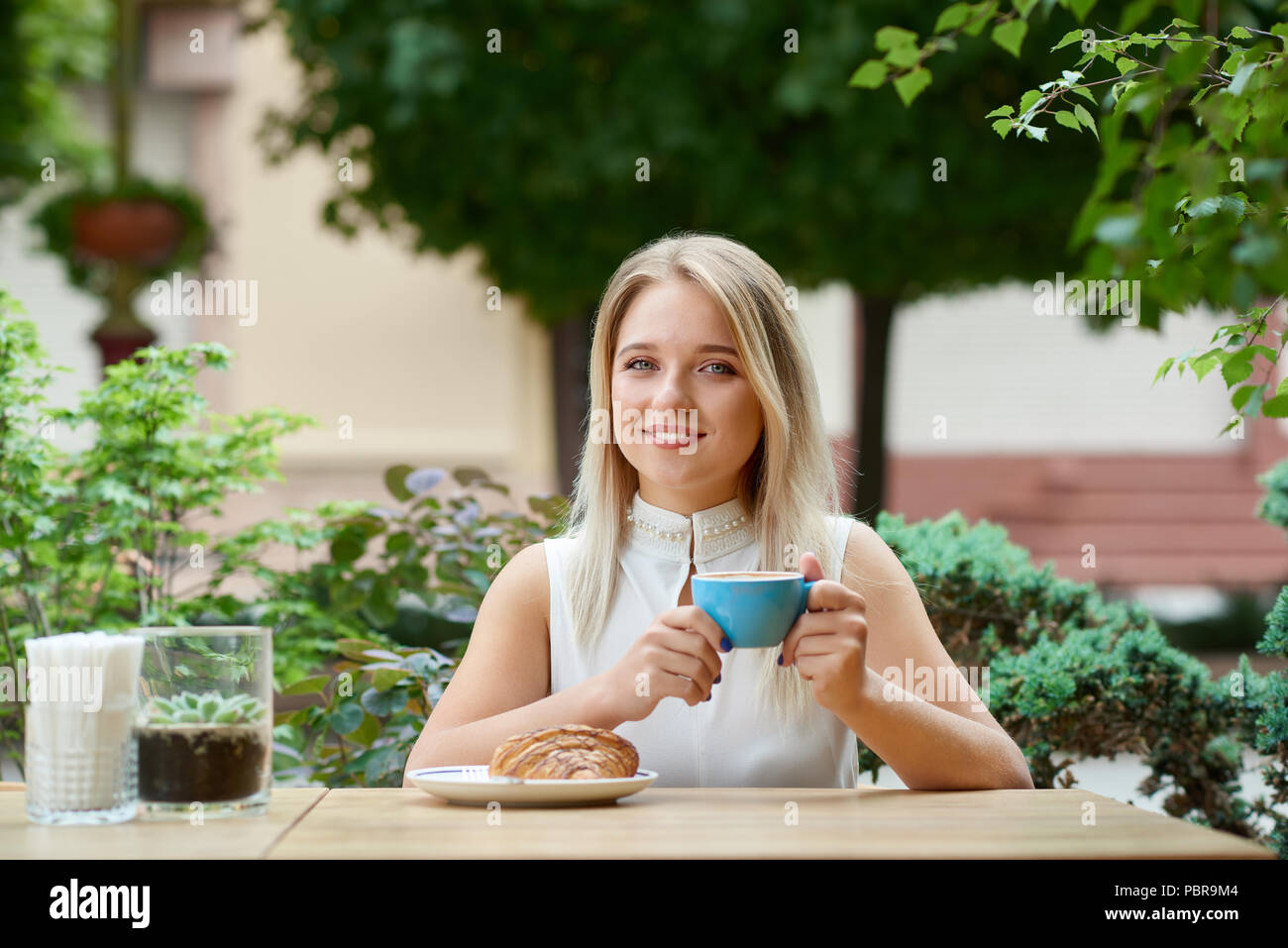 Jolie fille assise sur l'extérieur cafe lodge de boire du café, manger un  croissant. Avoir beau sourire, cheveux blonds, yeux bleus, portant chemise  blanche. Les plantes vertes et d'arbres sur fond de