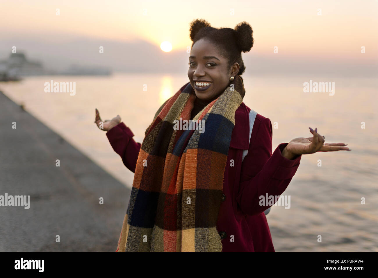 Portrait d'une femme africaine de 25 ans avec un manteau rouge et trame près du bord de l'eau avec un coucher de soleil. Banque D'Images