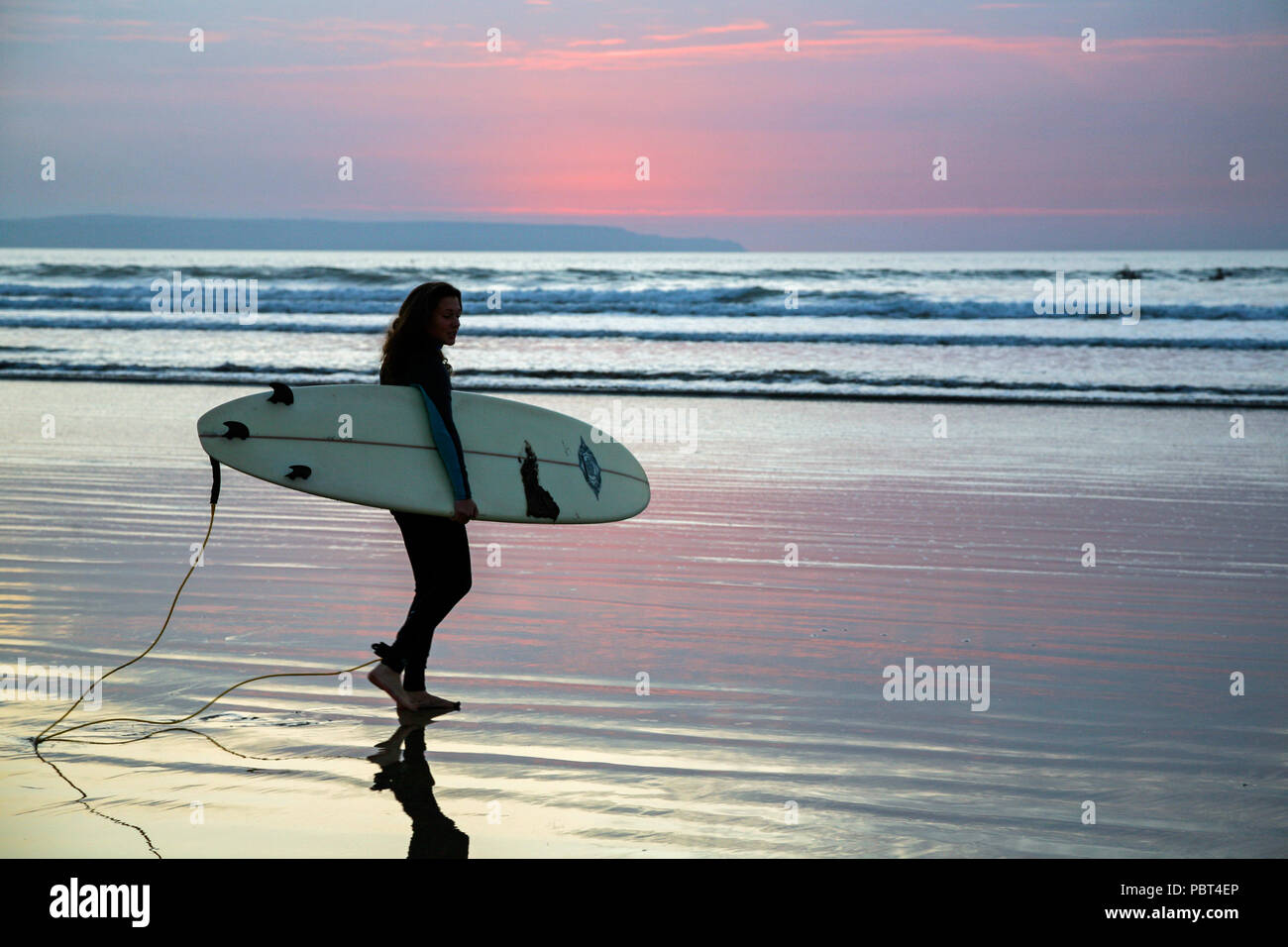 Jeune femme surfer carrying her surfboard comme elle partira dans la mer pour attraper une vague surfant à Westward Ho ! Beach, North Devon, Angleterre, Europe. Banque D'Images