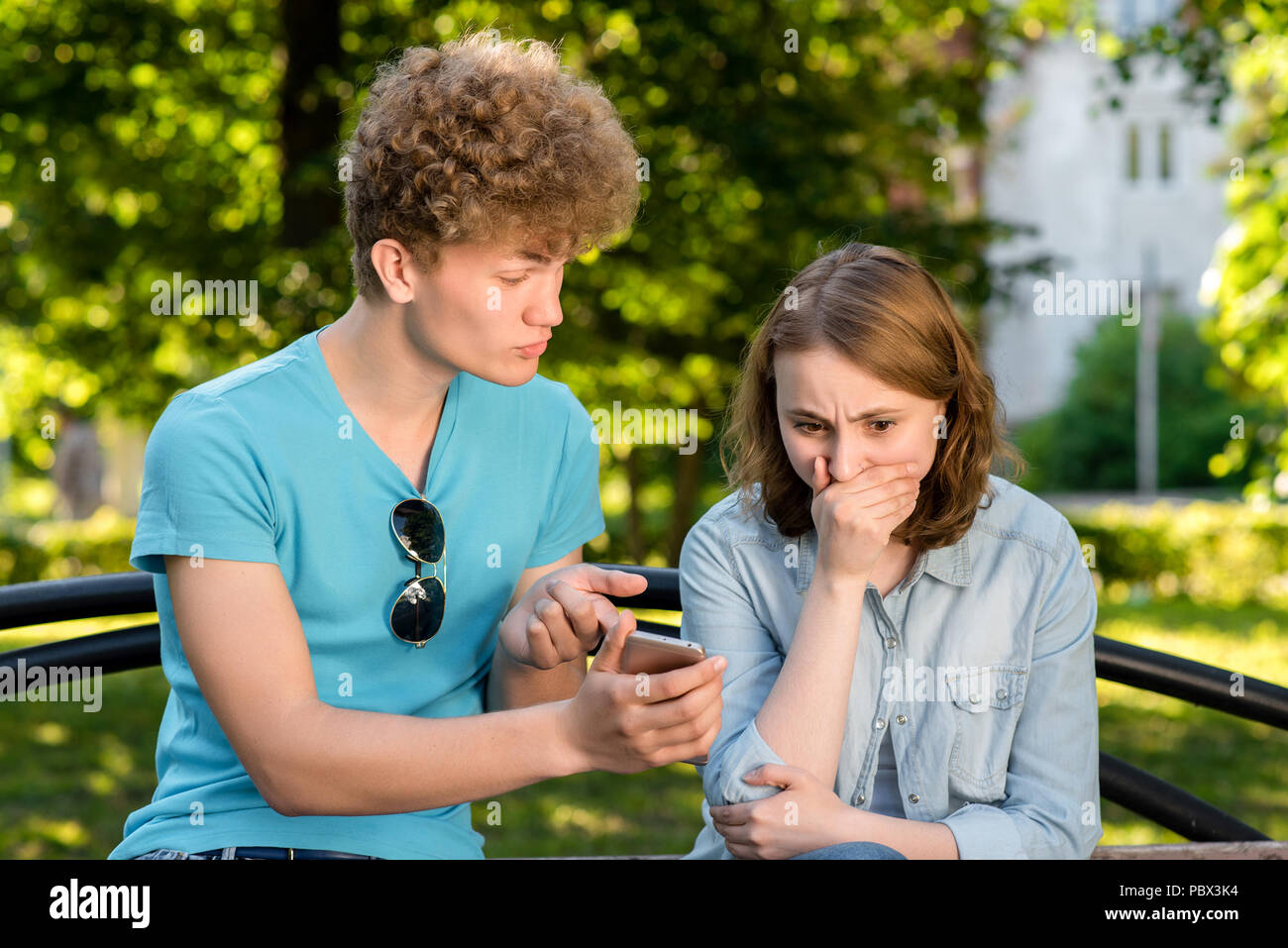 Un garçon avec une fille en été à l'extérieur. Un mec est main dans la main avec un smartphone pointant son doigt vers le téléphone. Les émotions de la méfiance. Le problème est en relation. Corrections dans la correspondance. Banque D'Images