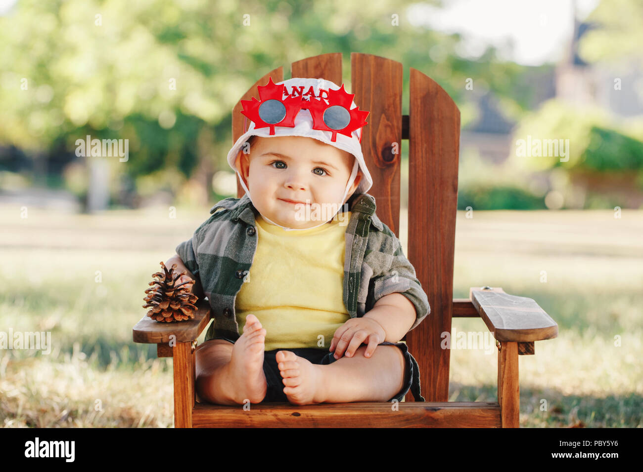 Portrait of little white Caucasian baby boy wearing hat Canadian maple leaf et drôles de lunettes. Enfant assis dans la chaire de Muskoka petit parc sur l'été Banque D'Images